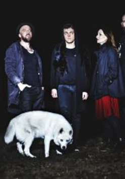 Zdjęcie przedstawia zespół muzyczny złożony z trzech mężczyzn i jednej kobiety oraz biały pies rasy husky na pierwszym planie.