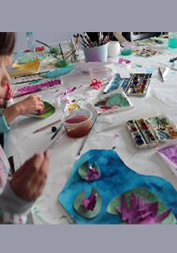 Stół z farbami i przyborami malarskimi, po lewej stronie dziewczynka z pędzelkiem