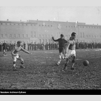 Zdjęcie czarno-białe: zawodnicy grający w piłkę nożną na boisku, na dalszym planie grupa kibiców
