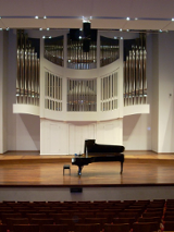 Zdjęcie przedstawia scenę auli Akademii Muzycznej w Poznaniu. Na środku sceny ustawiony jest fortepian. W tle widoczne piszczałki organów.