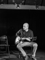 Zdjęcie mężczyzny siedzącego na krześle i grającego ma gitarze.