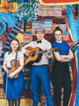 Zdjęcie trzyosobowego zespołu muzycznego (gitara, skrzypce i wokal). W tle namalowany kolorowy plan miasta.