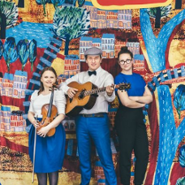 Zdjęcie trzyosobowego zespołu muzycznego (gitara, skrzypce i wokal). W tle namalowany kolorowy plan miasta.