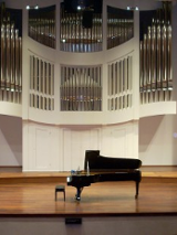 Na zdjęciu scena z fortepianem na środku. W tle piszczałki organów.