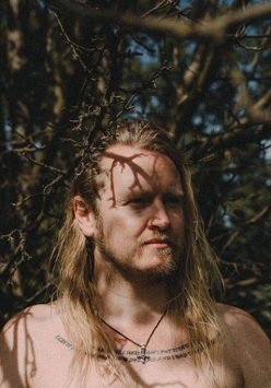 zdjęcie wykonawcy. Długowłosy mężczyzna z zarostem na tle bezlistnego drzewa.