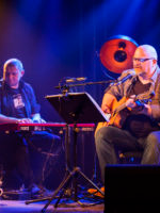 Zdjęcie przedstawia dwóch siedzących mężczyzn na scenie. Jeden z gitarą, drugi przy instrumencie klawiszowym.