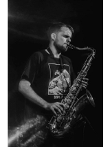 Czarno biała fotografia przedstawia profil młodego mężczyzny grającego na saksofonie.