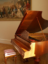 Na zdjęciu fortepian w kolorze drewna. Na ścianie fragment obrazu.