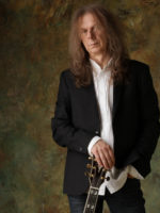 Zdjęcie Wojciecha Hoffmana. Długowłosy mężczyzna w czarnej marynarce i białej koszuli opiera prawą rękę na gryfie gitary.