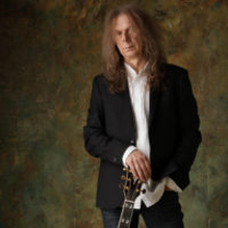 Zdjęcie Wojciecha Hoffmana. Długowłosy mężczyzna w czarnej marynarce i białej koszuli opiera prawą rękę na gryfie gitary.