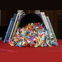 Stos figurek złożonych z origami na czerwonym stole. W tle stoi kilka książek.