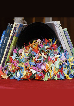 Stos figurek złożonych z origami na czerwonym stole. W tle stoi kilka książek.