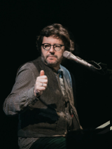 Na zdjęciu artysta siedzący przy fortepianie z mikrofonem przy twarzy.