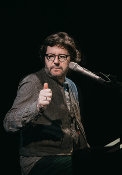 Na zdjęciu artysta siedzący przy fortepianie z mikrofonem przy twarzy.