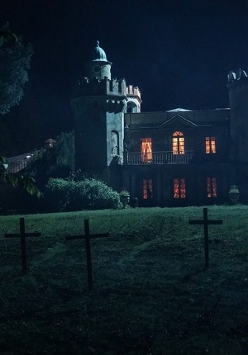 Kadr z filmu "Gniazdo zła" przedstawia gotycką posiadłość, stojącą na pagórku. Na zboczu pagórka krzyże. Jest ciemno i mroczno.