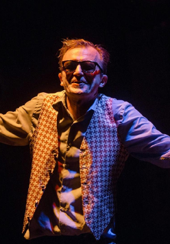 Na zdjęciu na czarnym tle, aktor w ciemnych okularach z rozłożonymi ramionami.