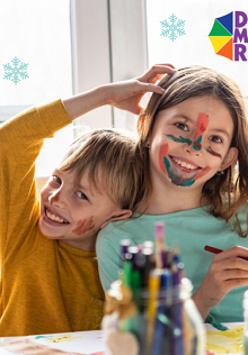 Na zdjęciu uśmiechnięte dzieci. Ich twarze są pomalowane w różnych kolroach. Na stole stoją kredki, ołówki i inne przybory do rysowania.