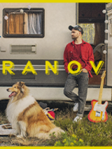 Na zdjęciu mężczyzna opierający się o przyczepę campingową. W otwartych drzwiach przyczepy stoi gitara. Przed przyczepą leży pies.