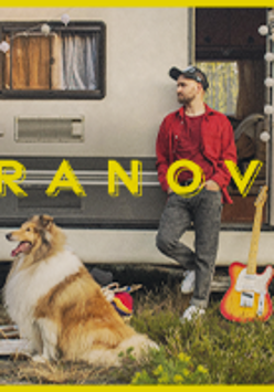 Na zdjęciu mężczyzna opierający się o przyczepę campingową. W otwartych drzwiach przyczepy stoi gitara. Przed przyczepą leży pies.