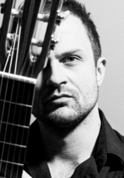 Na zdjęciu twarz mężczyzny. Prawą stronę twarzy przesłania mu gryf gitary.