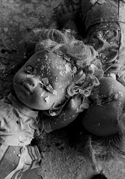 Czarno-białe zdjęcie przysypanych fragmentami pokruszonego tynku dwóch lalek, leżących na zniszczonym blacie stołu.