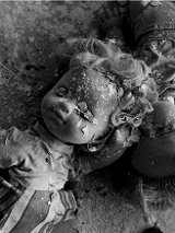 Czarno-białe zdjęcie przysypanych fragmentami pokruszonego tynku dwóch lalek, leżących na zniszczonym blacie stołu.