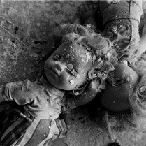 Czarno-białe zdjęcie przysypanych fragmentami pokruszonego tynku dwóch lalek, leżących na zniszczonym blacie stołu.