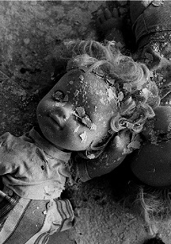 Czarno-białe zdjęcie przysypanych fragmentami pokruszonego tynku dwóch lalek, leżących na zniszczonym blacie stołu.