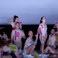 Grupa osób ubranych w cieliste stroje z kolorywami prostokątami. Ustawieni w różnych pozach.