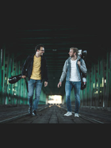 Dwaj muzycy idący po moście z instrumentami. Są zwróceni twarzami do siebie, uśmiechają się.