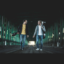 Dwaj muzycy idący po moście z instrumentami. Są zwróceni twarzami do siebie, uśmiechają się.