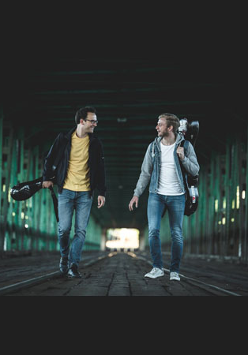 Dwaj muzycy idący po moście z instrumentami. Są zwróceni twarzami do siebie, uśmiechają się.