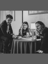 Czarno-biała fotografia. Na zdjęciu dwóch mężczyzn i kobieta. Siedzą przy stoliku kawiarnianym, w eleganckich strojach. Patrzą w obiektyw aparatu i się uśmiechają. Na stole filiżanki z kawą.