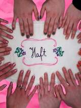 Sześć par dłoni kobiecych na różowym blacie. Na nim leży biały obrus z napisem "Haft".