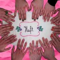 Sześć par dłoni kobiecych na różowym blacie. Na nim leży biały obrus z napisem "Haft".