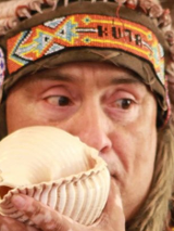 Mężczyzna w pióropuszu, w tradycyjnej indiańskiej kamizelce, dmucha w muszlę.