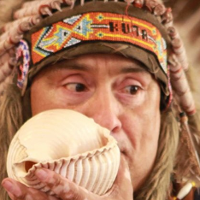 Mężczyzna w pióropuszu, w tradycyjnej indiańskiej kamizelce, dmucha w muszlę.
