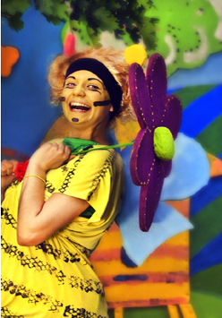 Na zdjęciu aktorka jako uśmiechnięta Pszczółka Maja trzymająca fioletowy kwiatek. Za nią wielobarwne tło we wzory roślinne.