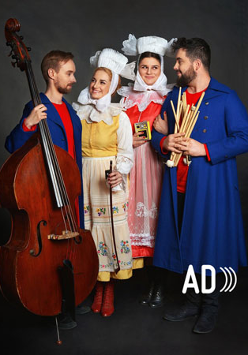 Na fotografii widać członków i członkinie zespołu Dagadana. Mężczyźni mają ubrane niebieskie, długie płaszcze i czerwone koszule pod nimi, kobiety zaś biało-żółte i biało-czerwone stroje stylizowane na ludowe. Muzyk po lewej stronie trzyma kontrabas. Artystki i artyści stoją obok siebie i spoglądają na siebie uśmiechnięci.