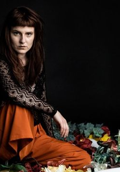 Na zdjęciu postać siedzącej artystki na czarnym tle.