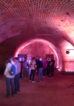 Grupa ludzi w podziemiach kaponiery, czytają plakaty rozstawione w pomieszczeniu.