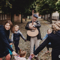 Grupa wesołych kobiet i dzieci tańczy na ulicy trzymając się za dłonie wokół uśmiechniętego mężczyzny przytulającego gitarę. Wzdłuż ulicy rosną drzewa, a w tle widoczne są budynki.