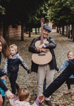Grupa wesołych kobiet i dzieci tańczy na ulicy trzymając się za dłonie wokół uśmiechniętego mężczyzny przytulającego gitarę. Wzdłuż ulicy rosną drzewa, a w tle widoczne są budynki.