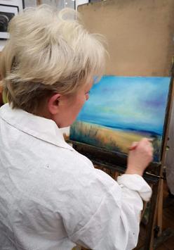 Starsza kobieta w białym fartuchu malująca obraz pędzlem na płótnie.