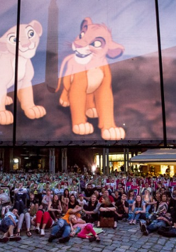 Grupa ludzi siedząca na ziemi pod ekranem z wyświetlonym filmem "Król lew".