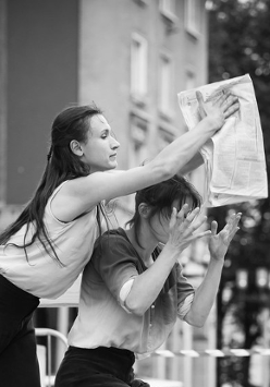 Zdjęcie czarno-białe. Dwie kobiety ujete w momencie wykonywania sceny. Jedna siedzi, a druga stoi nad nią i trzyma gazetę.