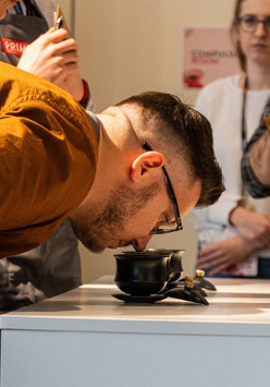 Zdjęcie przedstawiające mężczyznę schylonego i wąchającego napój w filiżance, obserwuje go grupa ludzi