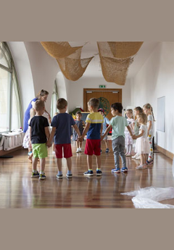 Na zdjęciu dzieci stoją na drewnianej podłodze w Bawialni w kole i trzymają się za ręce. Po lewej stronie widzimy prowadzącą zajęcia, a za nią duże okno w zielonej ramie.