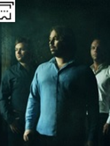 Zdjęcie przedstawia trzech muzyków w koszulach stojących na butelkowym tle.