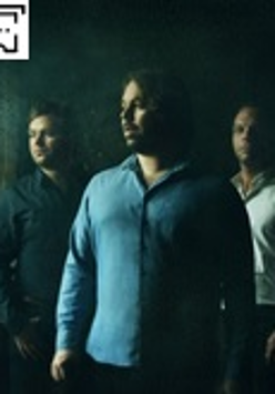 Zdjęcie przedstawia trzech muzyków w koszulach stojących na butelkowym tle.